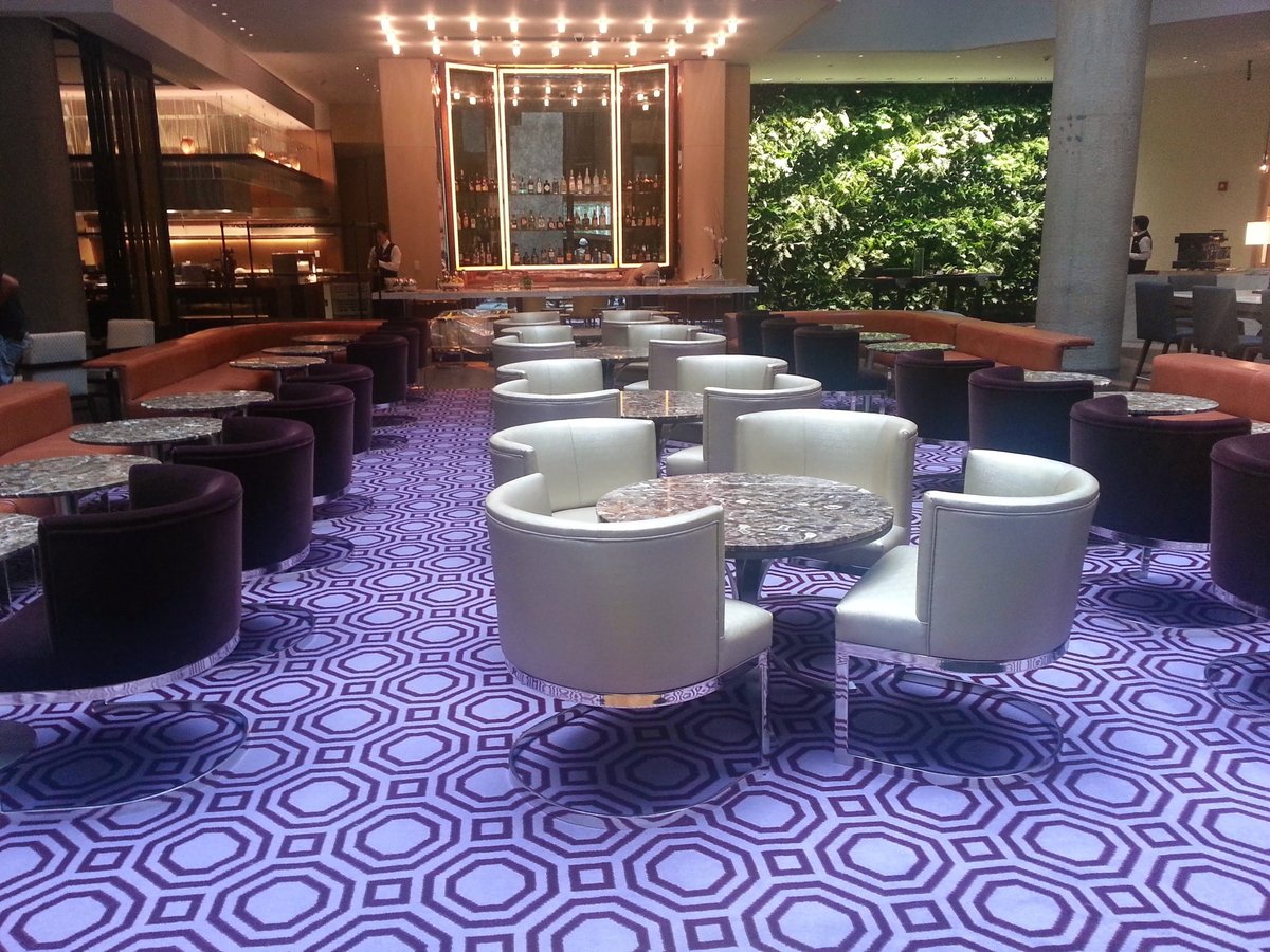 Lobby de hotel moderno con piso geométrico en morado, sillones cilíndricos blancos, mesas de mármol, barra con botellero iluminado y jardín vertical al fondo